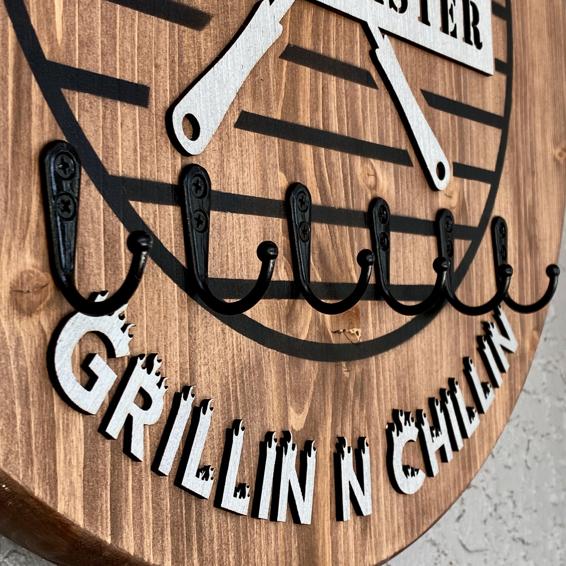 Close up of 6 metal hooks sturdy enough for holding bbq cooking utensils. HIgh-lighting the "Grillin n Chillin" letters at the bottom of the sign.